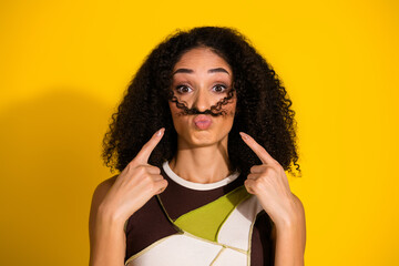 Playful young woman with curly hair mimicking moustache in front of a vibrant yellow background