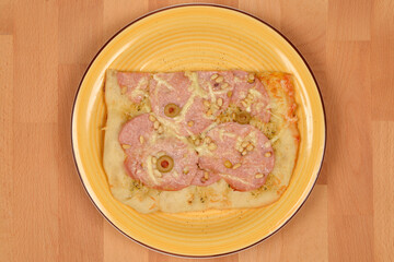 A piece of pizza with mortadella and pine nuts on a yellow plate, shot from above on a wooden...