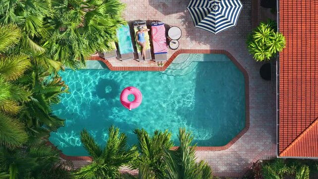 Aerial view of a tropical backyard pool where a woman relaxes on a sun lounger with colorful towels, a striped parasol nearby, and a pink flamingo float in the water, captured in cinematic slow motion