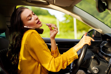 Woman driver multitasking, talking on cellphone and applying lipstick, while holding steering...