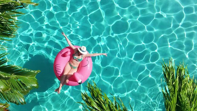 Aerial view pool with sparkling teal blue water with smiling woman floating on pink flamingo. Luxury travel concept of summer vacation in tropical Miami villa with lush palm trees on perfect sunny day