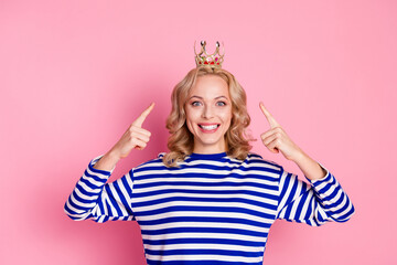 Confident young woman with a crown on a pink background gesturing proudly, wearing striped pullover