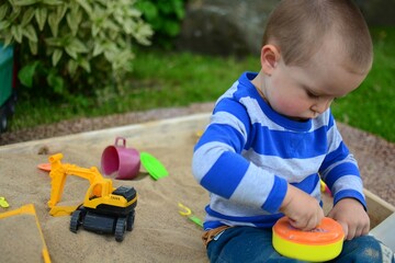 Toddler boy is playing in sandbox in a back yard . Child making sand meals with toy pan outside in summer time.
