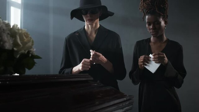 Low angle shot of two women in black, each holding tissue, standing somberly beside coffin at funeral