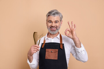 Smiling hairdresser with scissors and comb showing OK gesture on beige background