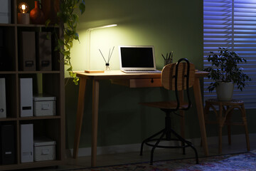 Comfortable workplace with laptop and lamp on wooden desk at home office in evening