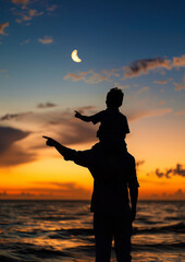 Fototapeta premium Father and Son Sunset Silhouette - A silhouette of a father and son watching the sunset by the ocean, with a crescent moon in the sky