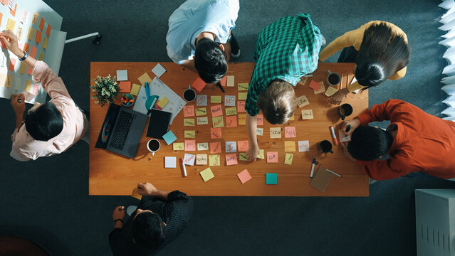 Top view of skilled business people discuss plan at meeting room while writing idea at sticky notes and stick at whiteboard. Group of diverse team working together to planning strategy. Convocation.