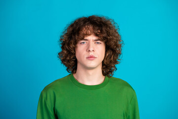 Young man with curly brown hair wearing green sweatshirt against blue wall background showcasing casual style and serious expression.