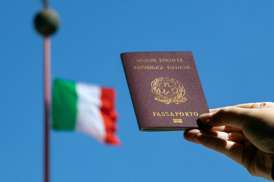 Italian citizenship - Hand holding a passport with Italian flag in the background against blue sky