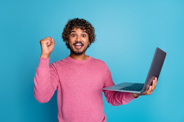 Excited Young Man Holding a Laptop in a Stylish Pink Sweater Against a Blue Background Making a...