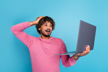 Young man in pink sweater holding a laptop and expressing surprise, isolated on a bright blue...