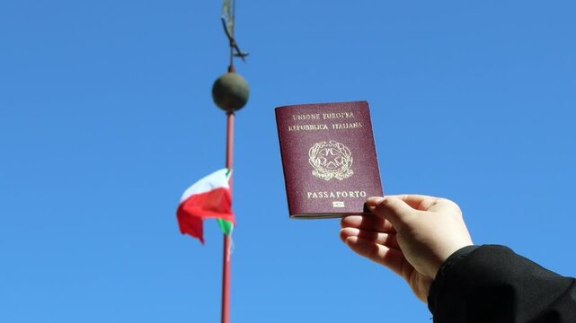 Italian citizenship - Hand holding an Italian passport with Italian flag in the background against blue sky