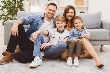Lovely Family Sitting On Floor Near Sofa Posing At Home, Smiling To Camera. Happy Parents And Children Concept