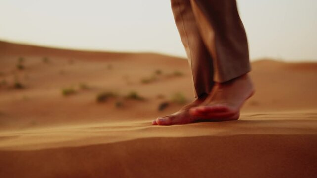 Bare male feet walking on sand dunes in the desert close-up. Slow motion: A close-up of a young man's feet walking on the sands in the desert during sunset. Human bare feet in trousers walks on sand.