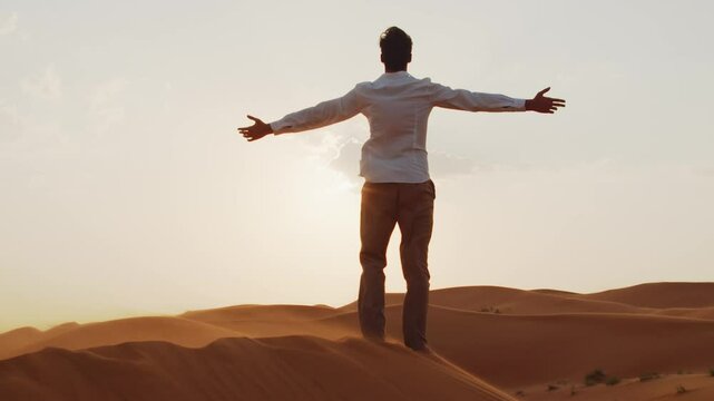 Young man in stylish clothes with raised hands up  in sand dunes in the desert at sunset. Back view of  young male person stands with hands up in the desert and looking to sun.  Freedom concept.