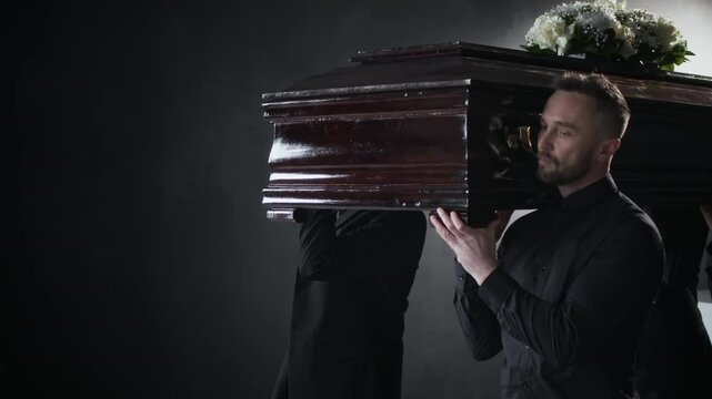 four men in black carrying polished, white-flowered, dark wooden coffin on their shoulders in somber funeral procession