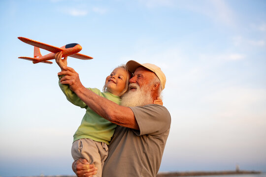 Joyful grandfather holding his happy blond grandson who is lifting a toy airplane into the sky, both enjoying playful time together under a clear blue sky.