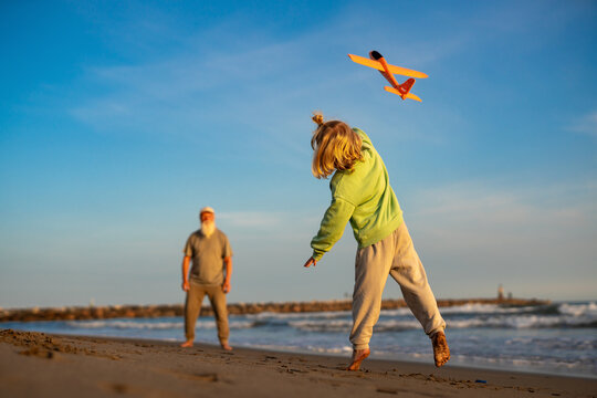 Barefoot young boy throwing a bright orange toy airplane on the beach at sunset, his grandfather standing in the background, joyful outdoor family activity at golden hour.