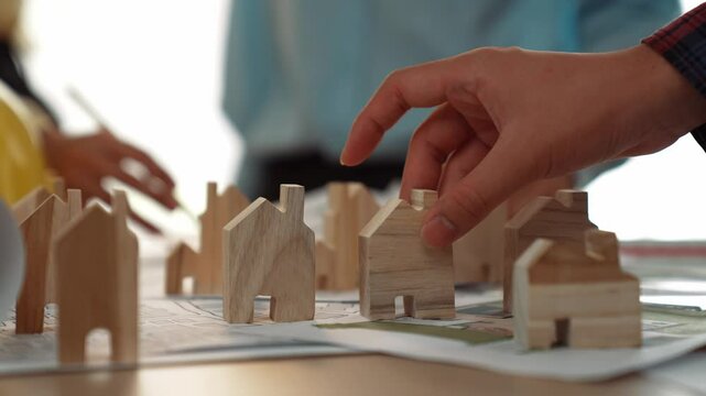 Architect and engineer discuss about building blueprint on table with architectural document, measurement tools scatter around at office, use wooden block for house positioning. Closeup. Delineation.