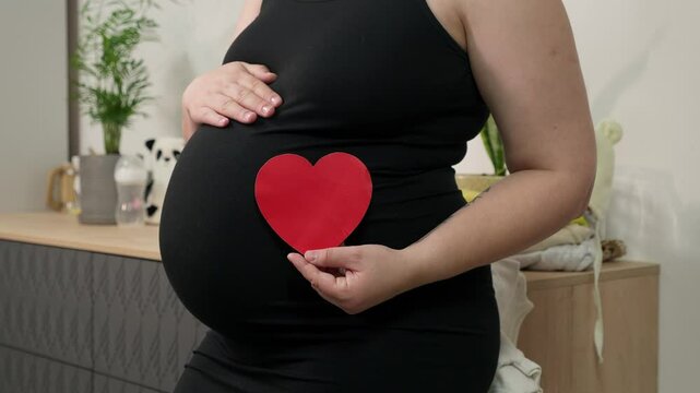Pregnant woman holding red heart shape and feel love for unborn baby in belly. Awaiting for baby. Concept of maternity and pregnancy care