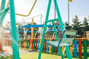 Empty swing seats at a vibrant colorful playground on a sunny day, with rainbow fence and play structures in the background