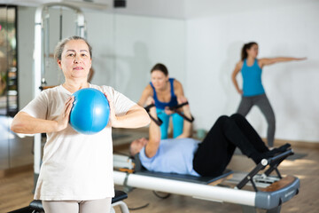 Willing elderly woman practicing pilates with ball in fitness hall during workout session. Persons...