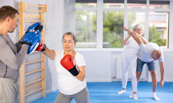Elderly lady, with help of teacher, improves boxing skills, learns to accurately and sharply strike opponent. Man trainer teaches effective protection during wrestling and dueling, martial arts.