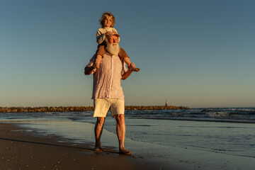 Happy grandfather carrying his young grandson on his shoulders while walking barefoot along the...