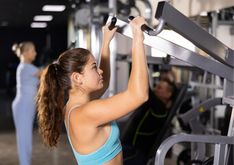 Fototapeta premium Young woman trains her arm muscles using a simulator in the gym