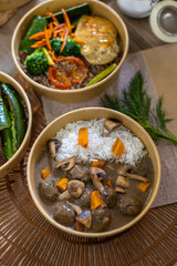 Close-up of a healthy meal with rice, meatballs, mushrooms, and pumpkin in an eco-friendly container. Placed on a rustic table with herbs and napkins. Vertical image, top view.