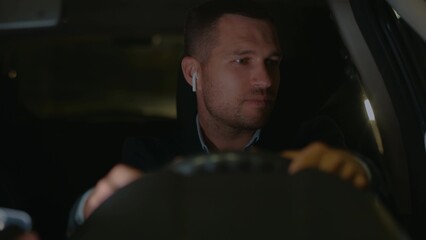 Joyful young businessman wearing suit and wireless earphones driving car in the evening. Caucasian...