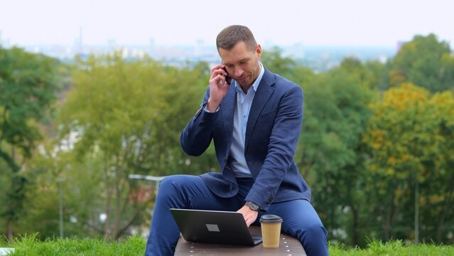 Camera approaching young businessman sitting outdoor chatting on smartphone and using laptop while drinking coffee. City urban style. Caucasian handsome male talking on cellphone
