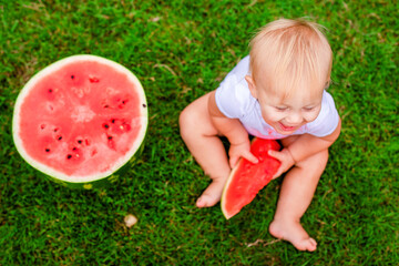 Blonde toddler biting into juicy watermelon while sitting barefoot on lawn