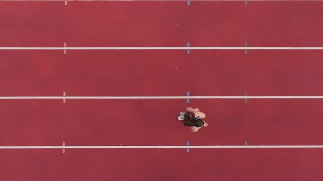 Top down view aerial drone shot of a professional female athlete sprinting on a large stadium athletics track. Dynamic slow motion cinematic footage captures her power, speed, and determination in