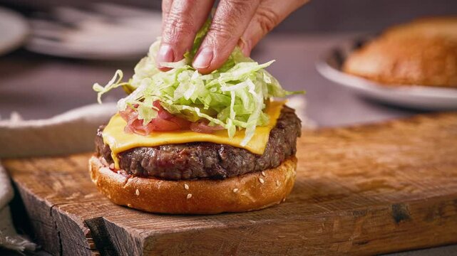 cook put chopped lettuce on grilled burger meat for making delicious cheeseburger on wooden cutting board, food closeup