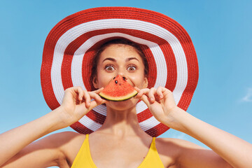 Young woman with a playful expression holding a slice of watermelon in front of her mouth while...