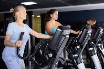 Focused aged woman leading healthy active lifestyle doing cardio workout on elliptical machine in...