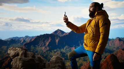 Male content creator recording a video surrounded by mountain peaks in the clouds, climbing a mountain range hill and live streaming for his social media audience. Adventurous spirit. Camera A.