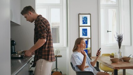 Husband preparing weekend breakfast for family. Relaxed woman browsing mobile