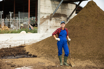 Female farmer with shovel poses against pile of dried and crushed cow dung in the backyard of a farm