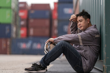 A tired logistics worker in a gray uniform sits on the ground at a busy shipping container yard,...