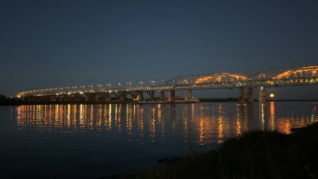 A bridge over a river at night with lights on it. The bridge is lit up and the water is calm
