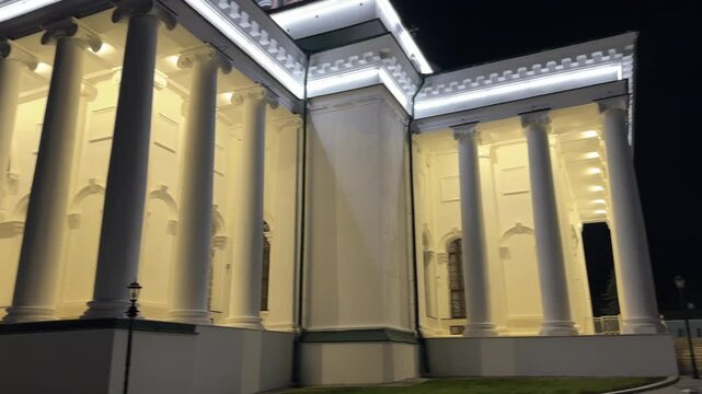 A large white building with a tall white pillar in front of it. The building is lit up at night