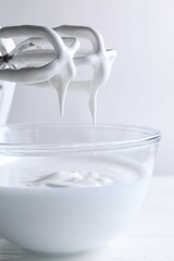 Hand mixer with whisks and bowl of whipped cream on white wooden table, closeup