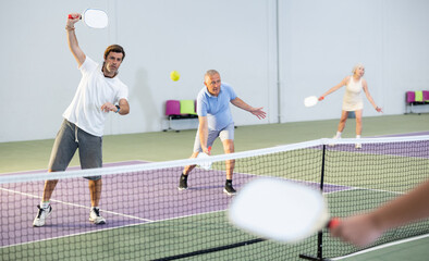 Obraz premium Portrait of concentrated adult pickleball player preparing to strike and return ball to opponents field during doubles match in team with older partner on indoor court