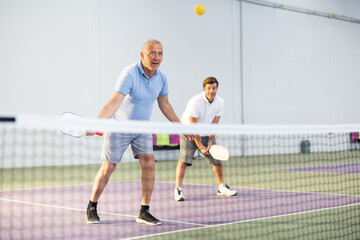 Portrait of sporty senior man playing doubles pickleball with male partner on indoor court, ready to hit ball. Sport and active lifestyle concept..