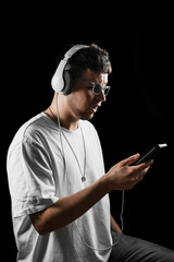 Young man in white t-shirt and headphones looking at smartphone, listening to music or podcast. Isolated on black background. Modern digital lifestyle.