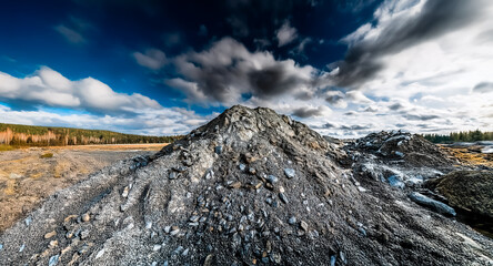 Naklejka premium A large mound of grey rocks dominates the foreground under a sky filled with dramatic blue and white clouds. A natural landscape with trees is visible in the background.