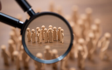 A magnifying glass highlights a focused group of wooden figures amidst a larger, blurred crowd on a wooden surface, emphasizing selection and attention to detail.